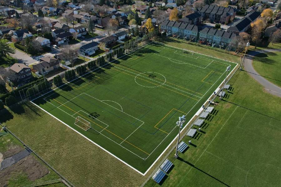 Aerial view of a rectangular artificial turf soccer field with markings, surrounded by houses and seating stands