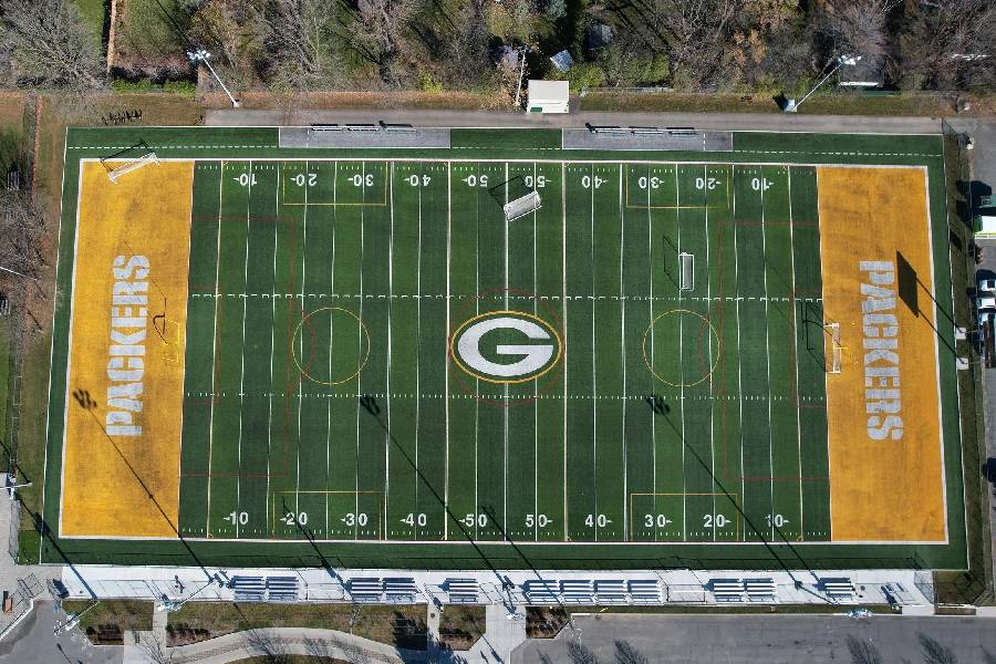Aerial top view of artificial turf football field with Packers branding, yard lines, and surrounding seating area