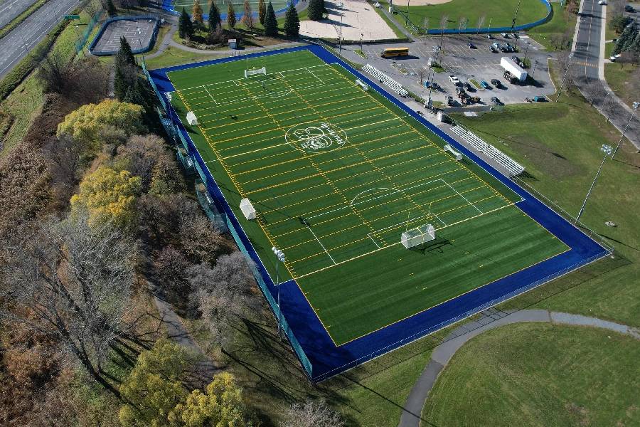Aerial drone view of a multi-sport artificial turf field with soccer goals, blue running track, parking area, and surrounding trees