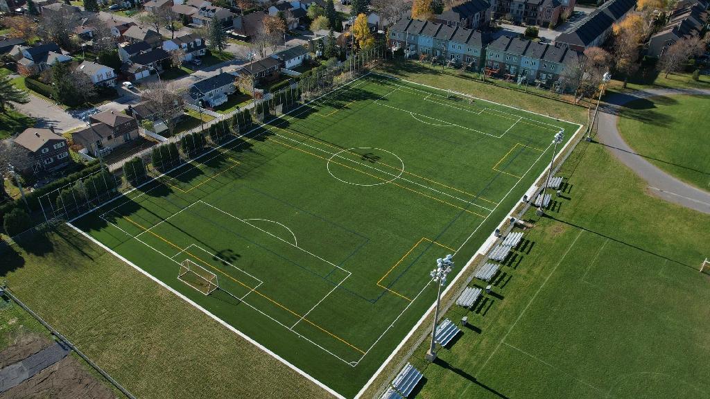 Aerial view of a community synthetic soccer field with marked playing lines, goalposts, spectator benches, lighting poles, and surrounding residential neighborhood.