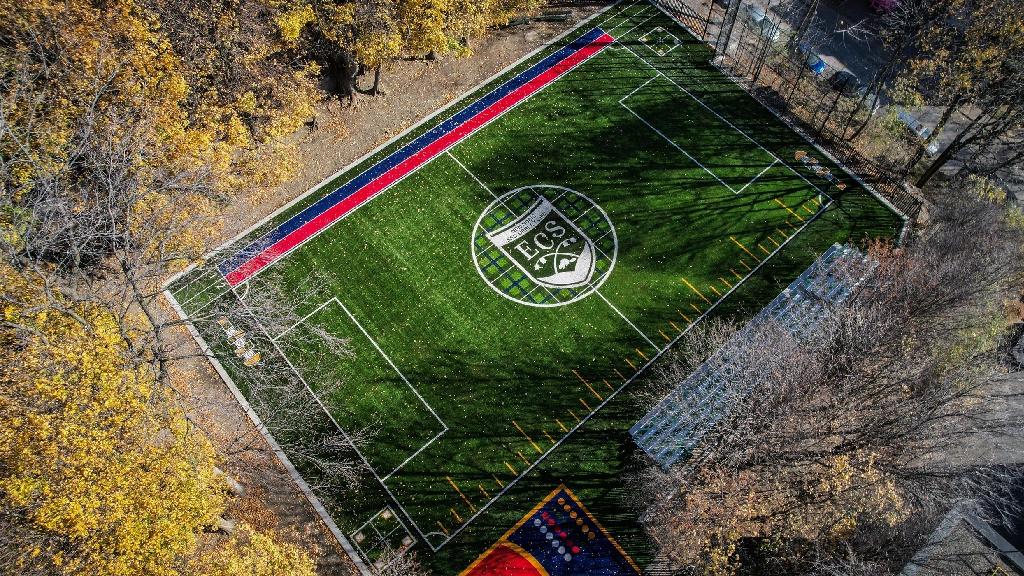 Aerial view of ECS School Westmount synthetic sports field featuring soccer markings, school logo at center, colorful boundary design, bleachers, and surrounding autumn trees.
