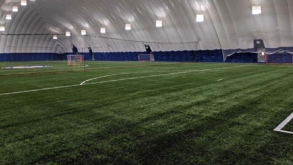 Large indoor dome facility featuring synthetic turf sports field, bright overhead lighting, marked playing lines, and spacious enclosed training environment.