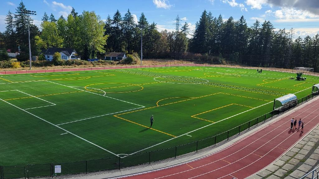 Wide view of a multi-sport synthetic turf field with soccer and football markings, surrounding running track, stadium lights, and forested landscape backdrop