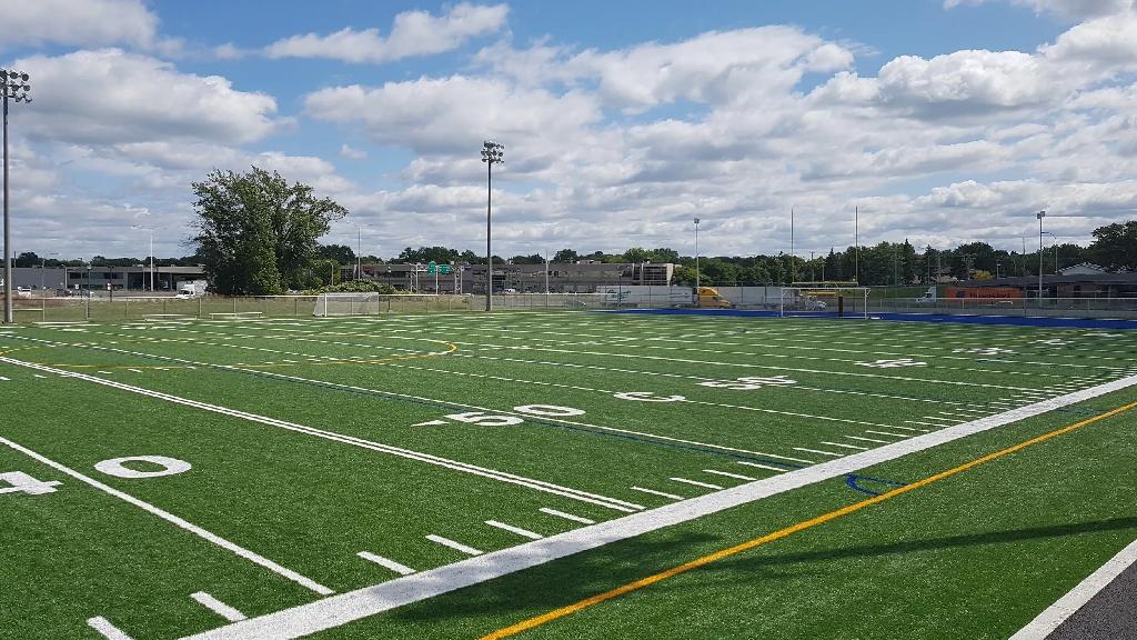 Wide daytime view of an outdoor synthetic football field with marked yard lines, stadium lights, soccer goals, and open sky with scattered clouds.