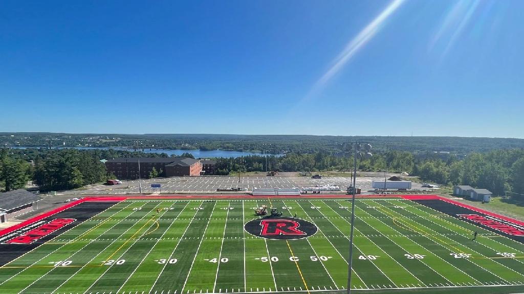 Wide view of Reds University synthetic turf football field with marked yard lines, large center logo, red end zones, stadium lighting, and scenic landscape in the background.
