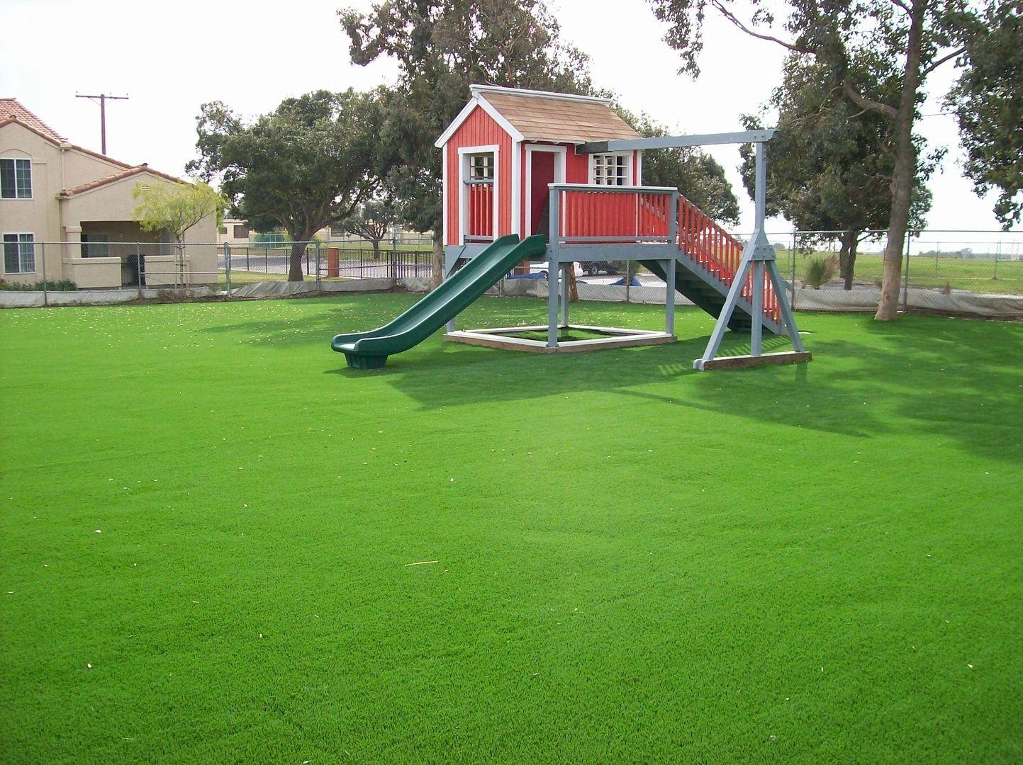 Residential backyard with artificial grass lawn, elevated children’s playhouse, slide, and spacious safe play area