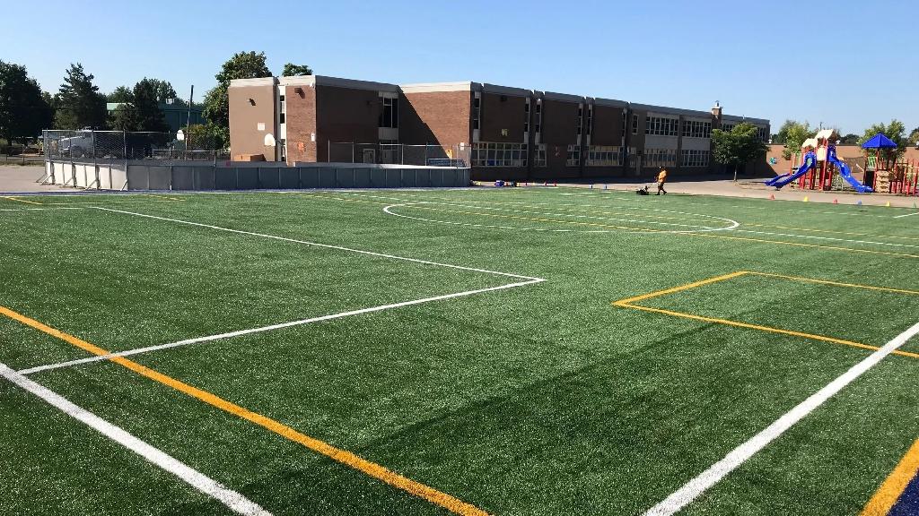 Synthetic multi-sport field at a school campus with marked soccer lines, playground area, school building, and bright outdoor setting.