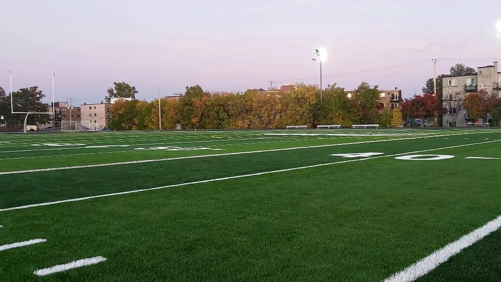 Wide evening view of a synthetic football field with bright stadium lights, marked yard lines, goalposts, and residential neighborhood backdrop.